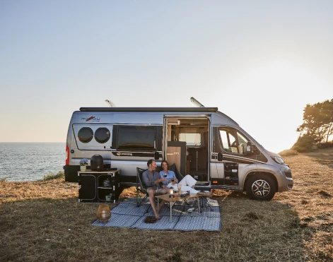 Camping-Van mit Paar und Camping-Möbeln vor einem See